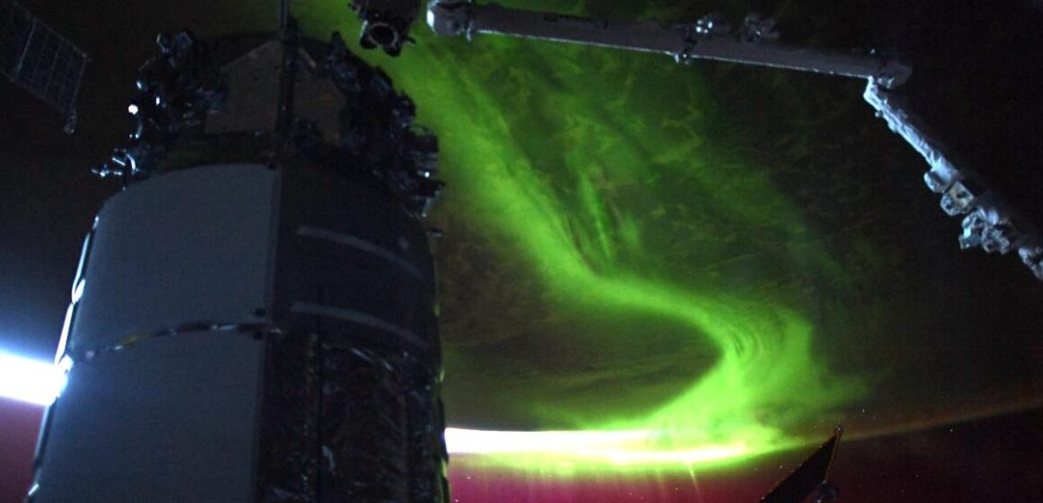 an aurora reaching into space from earth. a robot arm and a module are in the front, in shadow